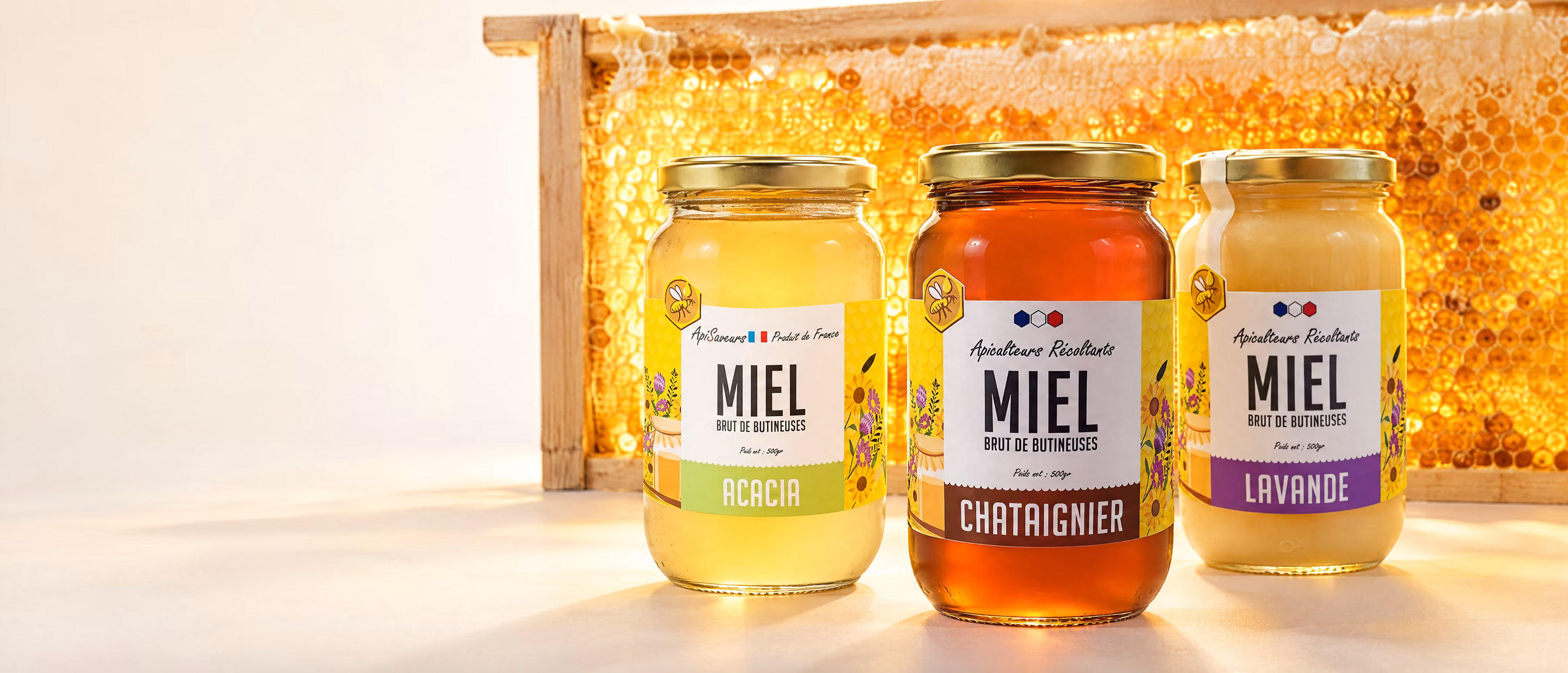 Trois pots de miel de France, acacia, châtaignier et lavande, devant un cadre de rayon de miel éclairé par l’arrière